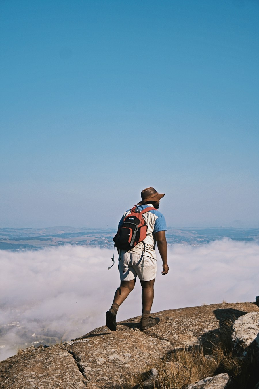 Hiker with a hat and backpack on a granite dome, facing a sea of clouds above the hills