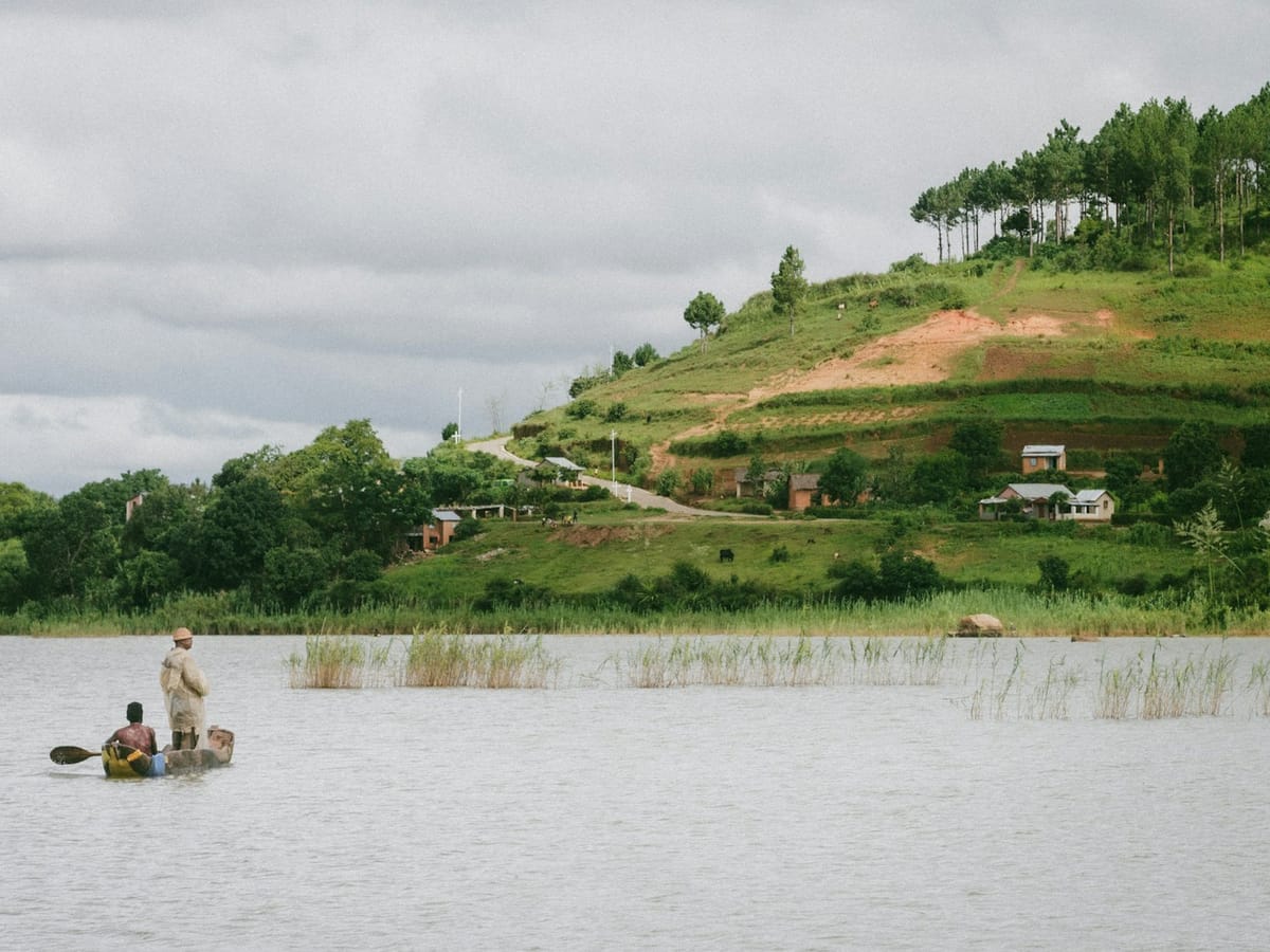 Two people in a dugout canoe on Lake Itasy near Ampefy, facing a grassy hill with houses and pine trees.
