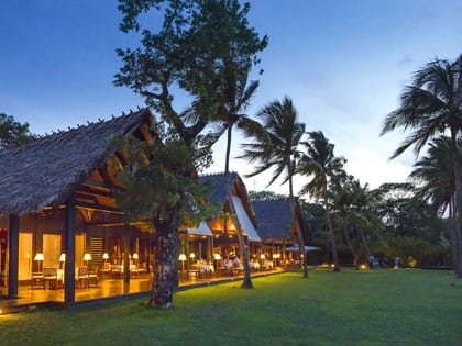 Thatched wooden buildings of the Anjajavy hotel lit up at dusk, beside a lawn and palm trees.