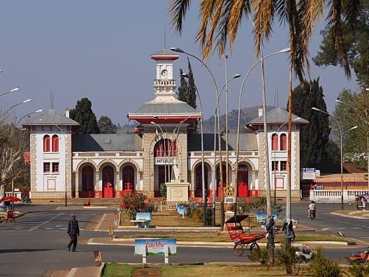 White and red façade of Antsirabe railway station with its small clock tower, facing a square lined with lamp posts.