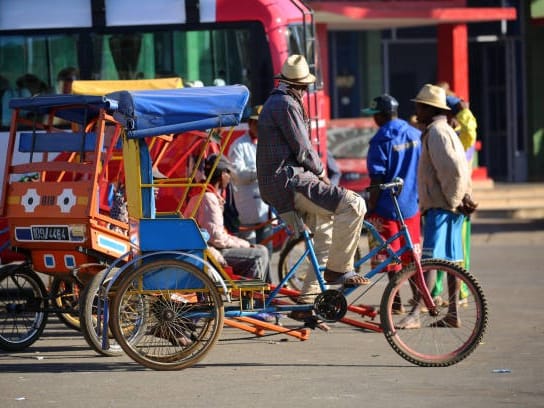 Brightly painted cycle rickshaws with a hatted driver and passengers in an Antsirabe street, in front of a red bus.