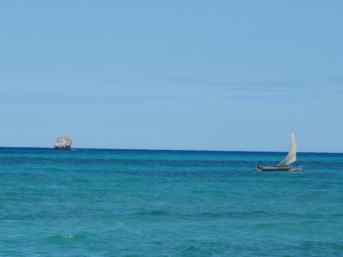 A canoe with a triangular sail skimming across the turquoise sea off Nosy Iranja, a rocky islet on the horizon.