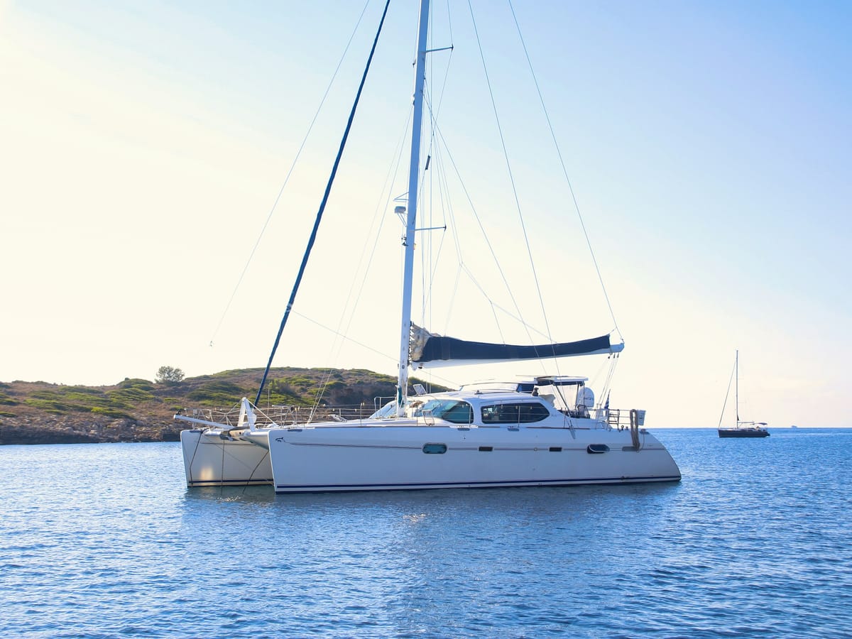 A white cruising catamaran lying at anchor on a calm sea near a low, scrub-covered headland, with a yacht in the distance.