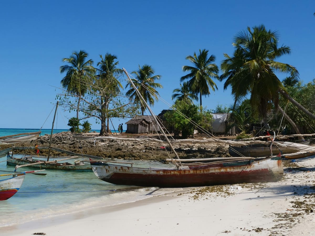 Wooden outrigger canoes pulled up on a white sand beach, sails furled, in front of the huts of a fishing village shaded by coconut palms, on Nosy Iranja.