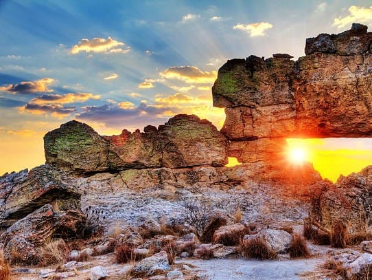 Low sun shining through the Isalo Window, a natural opening in a sandstone rock, beneath a streaked cloudy sky.