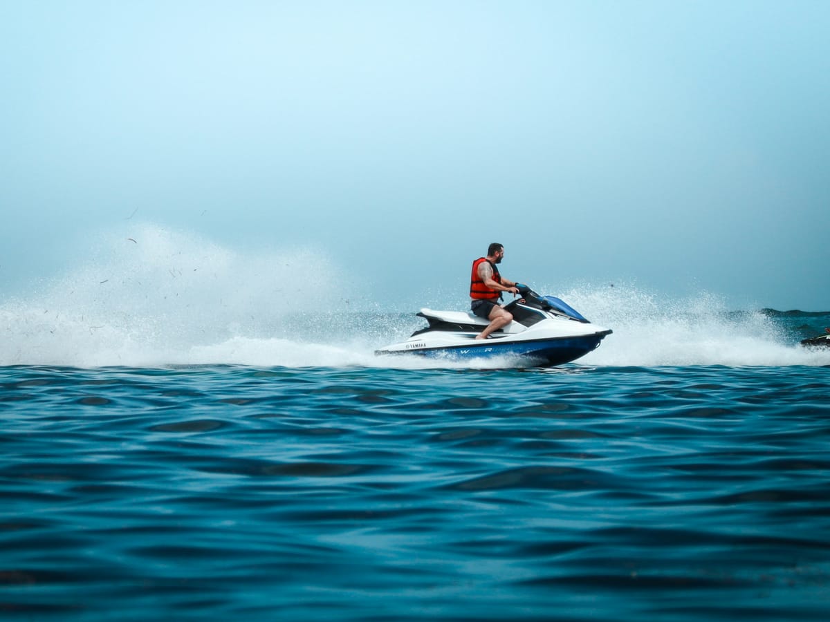 A rider in an orange life jacket on a white and blue jet ski, throwing up spray across a deep blue sea.