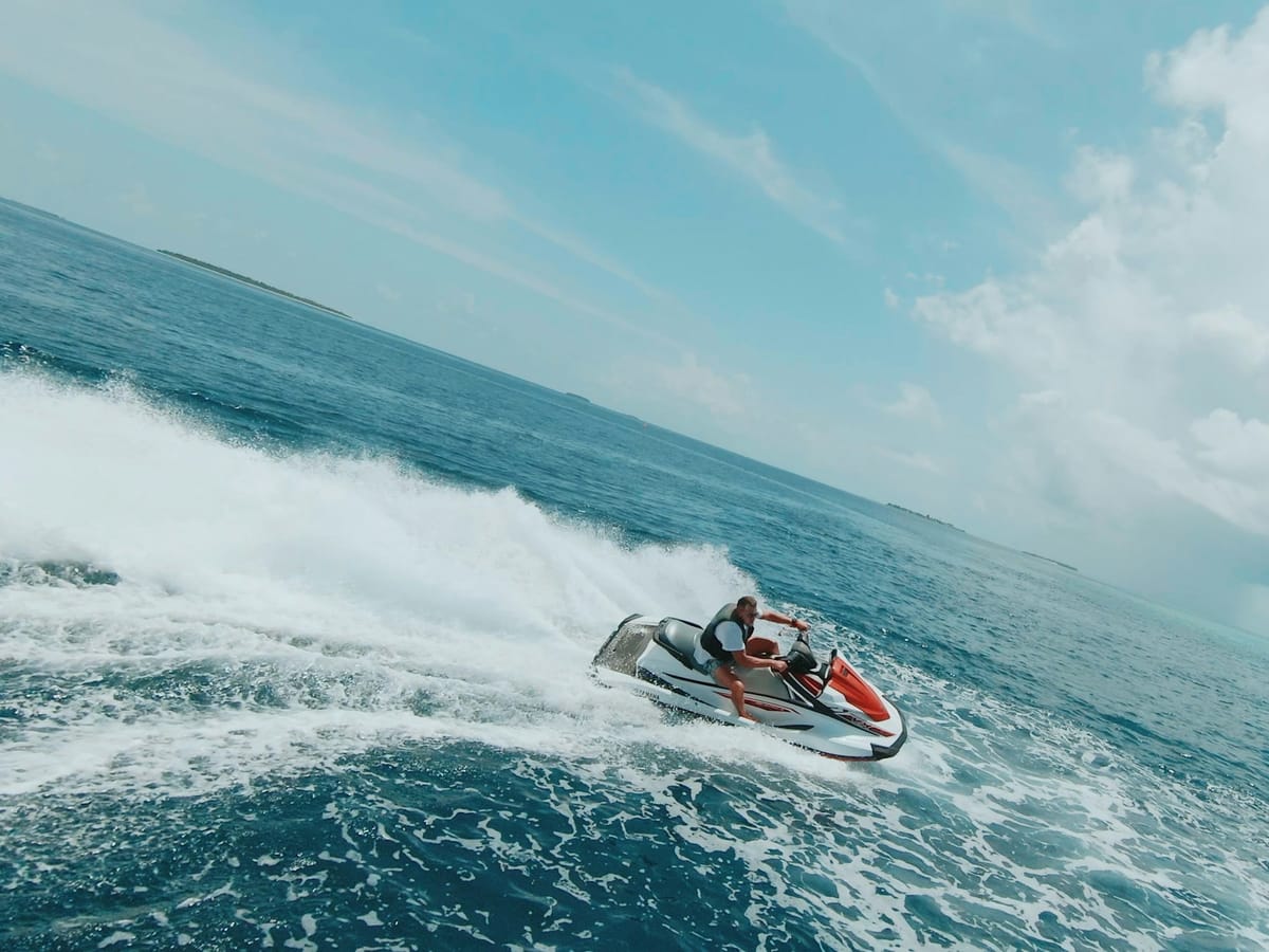 A rider in a life jacket banking on a white and red jet ski, leaving a wide trail of foam across a blue sea beneath white clouds.