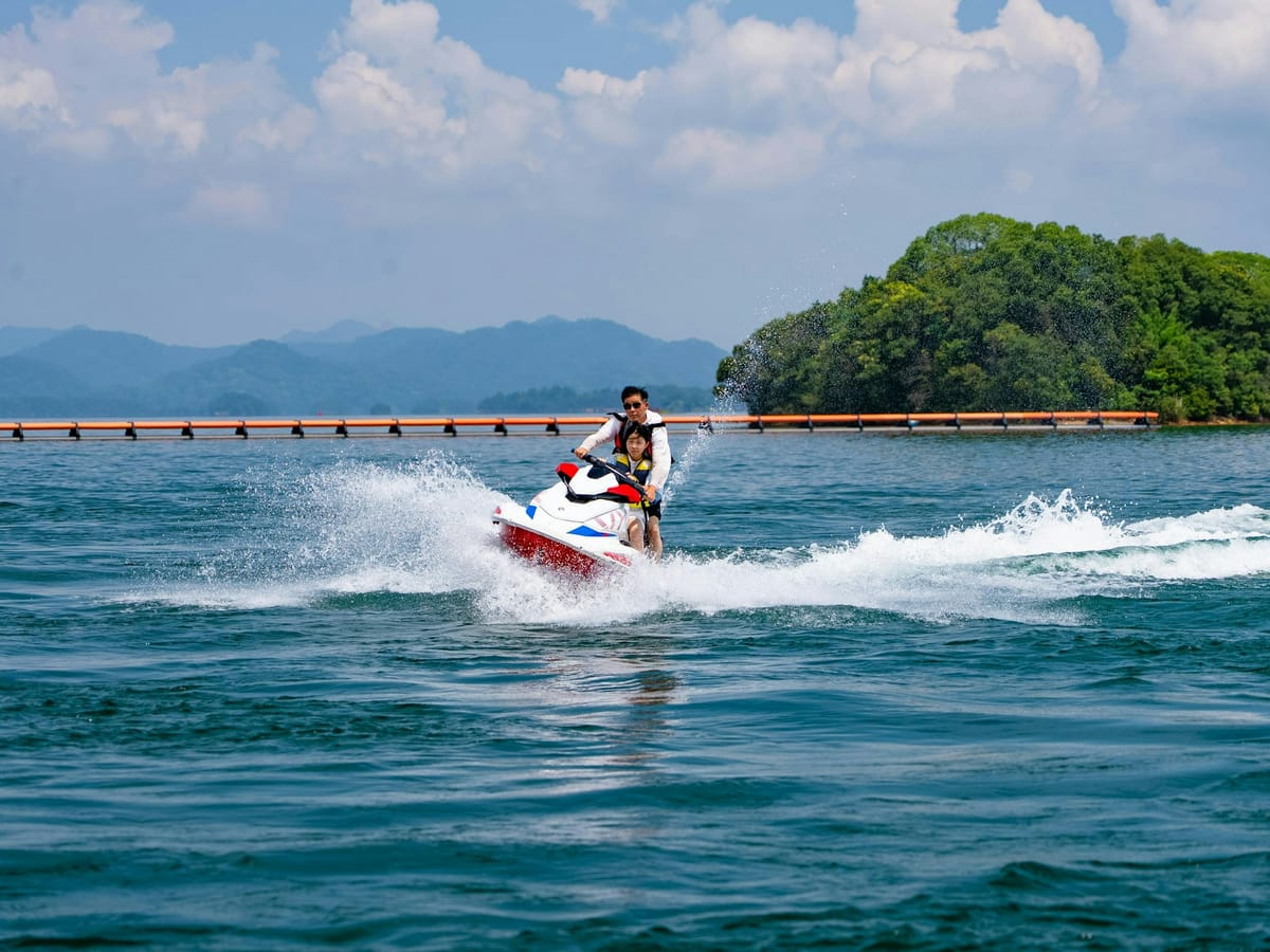 An adult and a child in life jackets on a white and red jet ski, speeding across a lake below wooded hills.