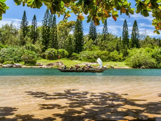 Sailing canoe carrying passengers across clear water lined with pine trees, near Lokaro Bay.
