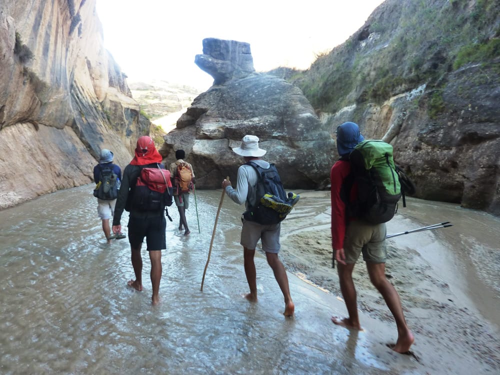 Hikers with rucksacks walking barefoot through a shallow river at the bottom of a canyon in the Makay.