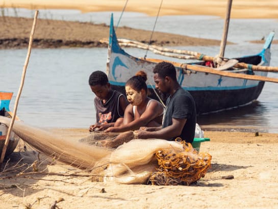 Three fishers sitting on the sand mend a net beside outrigger canoes on the Morondava coast.