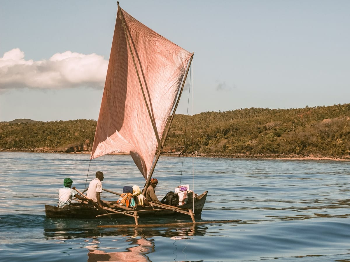 Outrigger pirogue with a pink sail carrying several passengers past a wooded coastline in the waters of Nosy Be.