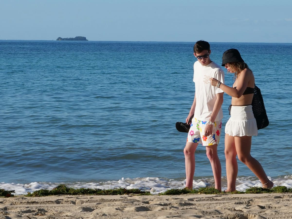 A young couple walking along the water's edge on a Nosy Be beach, a wooded islet on the horizon.
