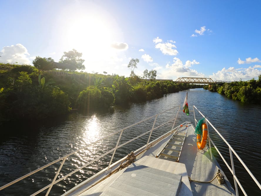 Bow of a boat flying the Malagasy flag on the Pangalanes Canal, lined with greenery, a steel bridge beyond.