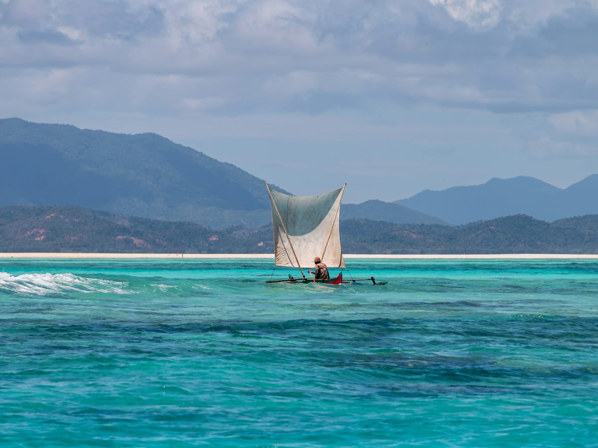 A lone fisherman in an outrigger pirogue with a white sail on a turquoise lagoon, with wooded hills behind, in Madagascar.