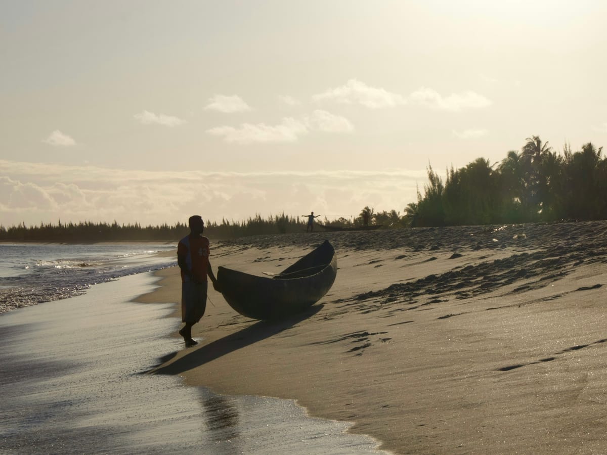 A fisherman silhouetted as he drags his dugout pirogue up the sand at the water's edge in the golden evening light, on a beach in Madagascar.