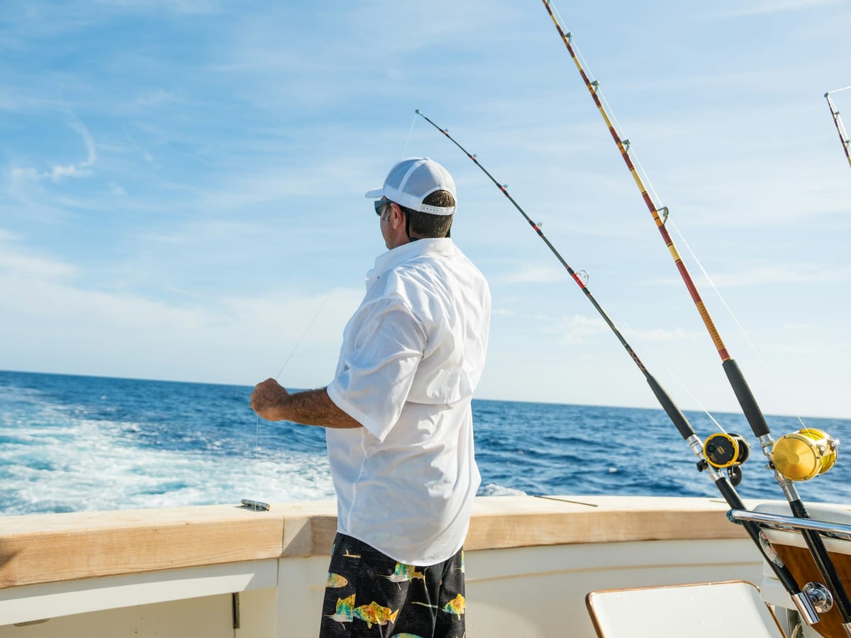 An angler in a white shirt at the stern of a big-game fishing boat, rods and reels set, looking out to the open sea.