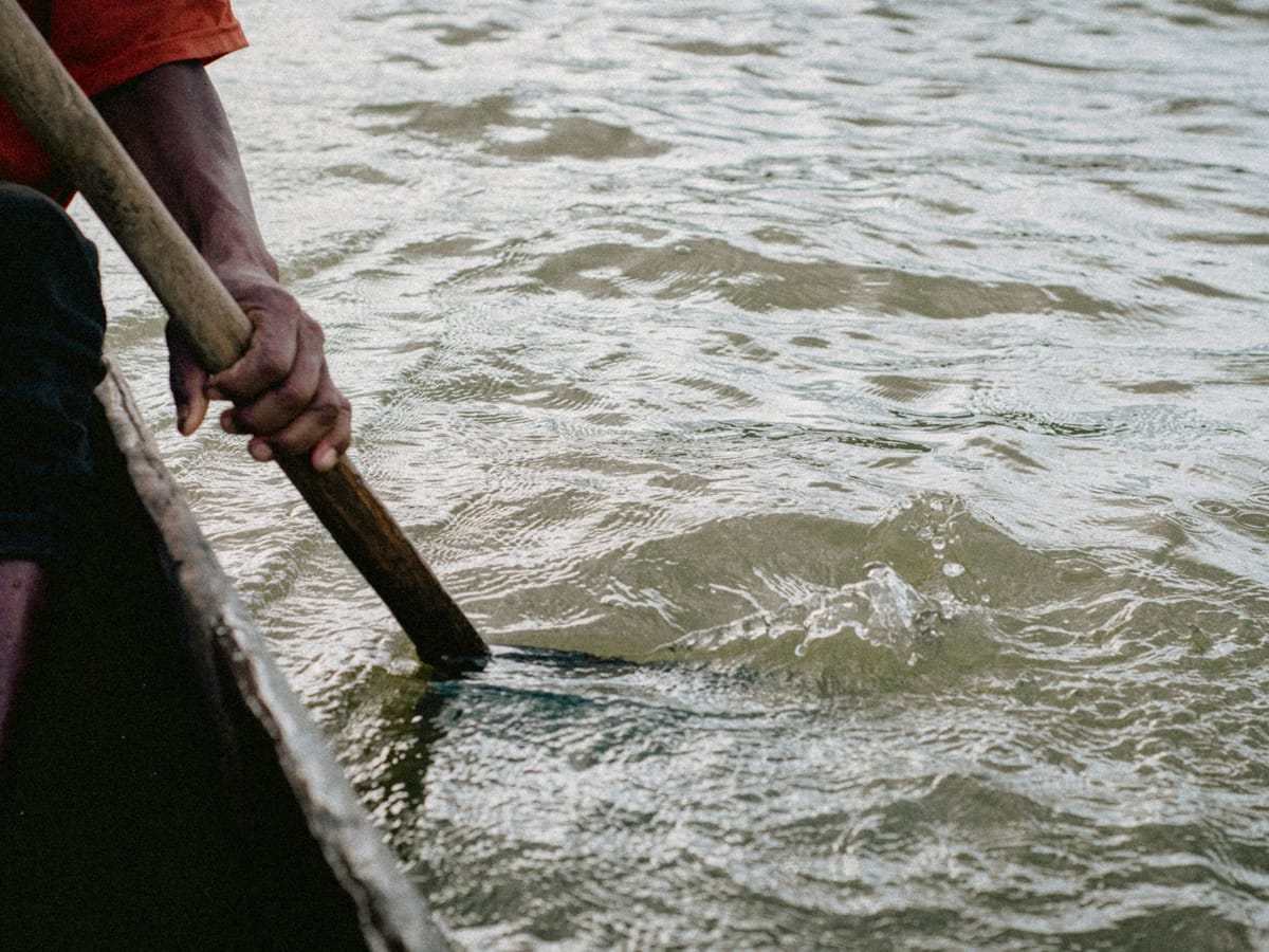 A boatman's hand dipping a wooden paddle into calm water from the side of a dugout canoe.