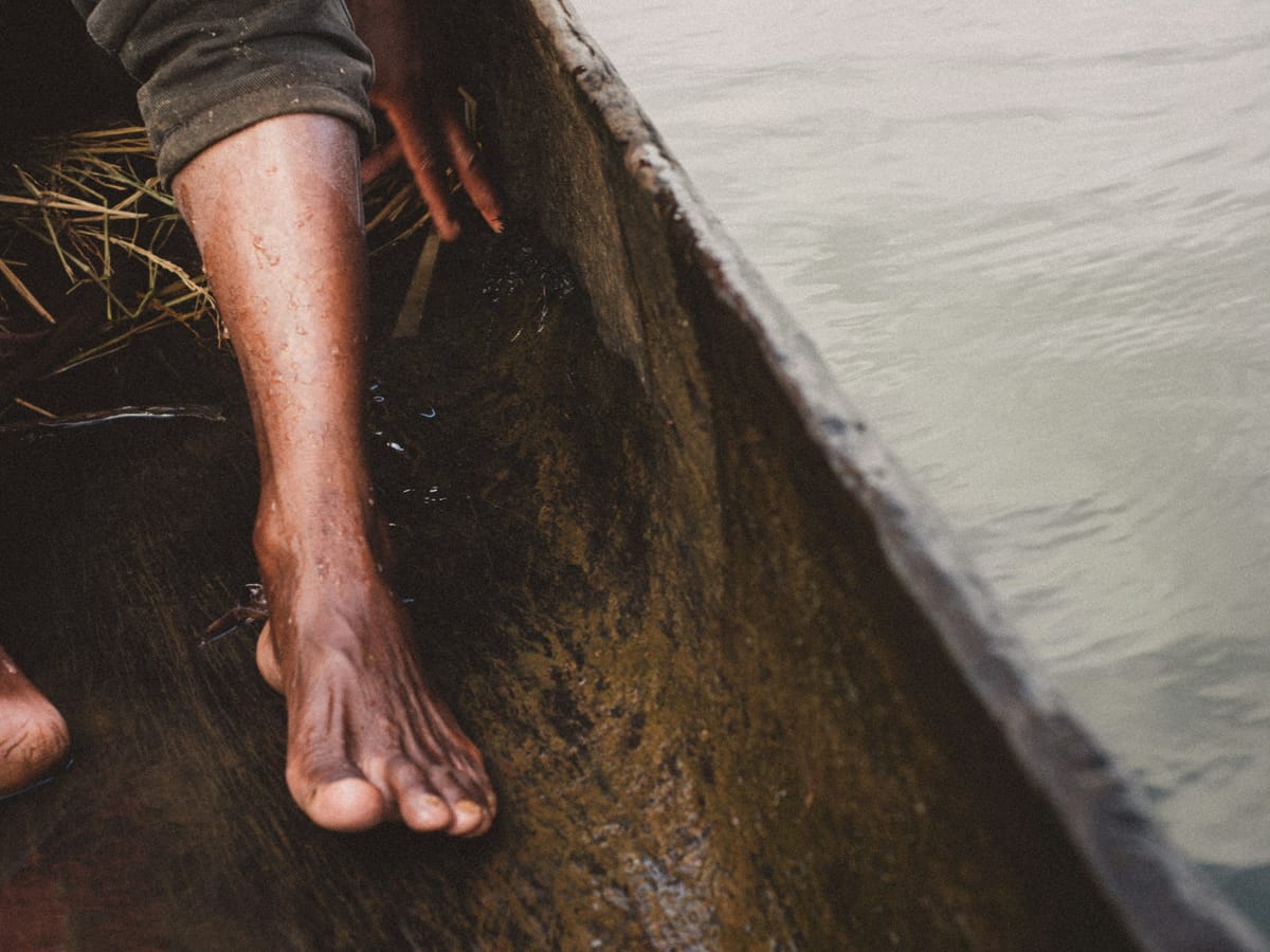 A boatman's bare feet resting in the bottom of a dugout canoe, its hull hollowed from a single tree trunk skimming the calm water.