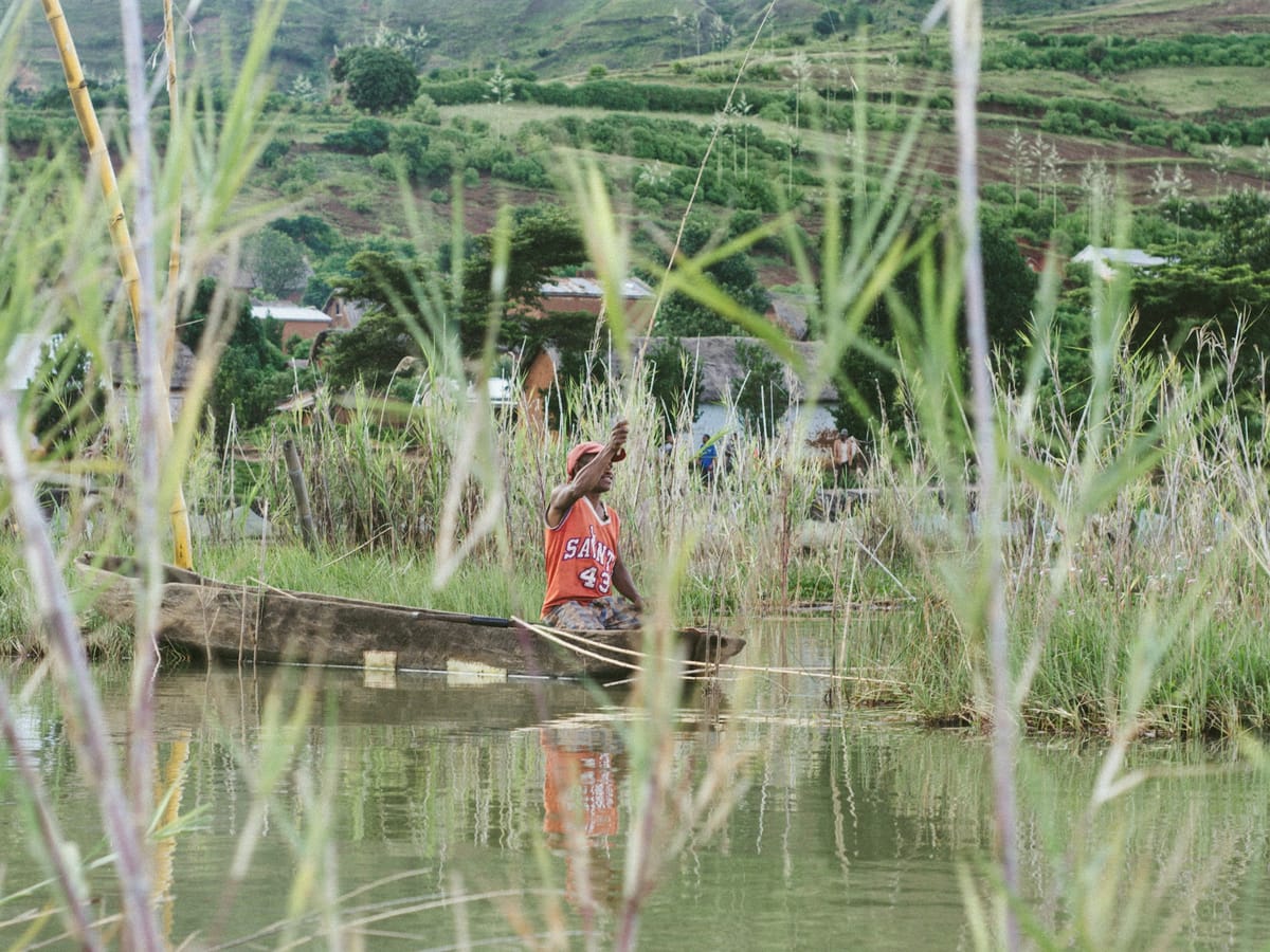 A man seated in his dugout among the reeds, in front of a village and terraced hillsides.