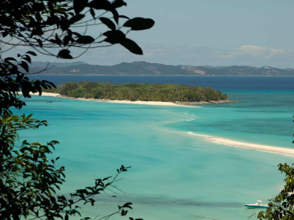 The turquoise lagoon of Nosy Iranja seen from high ground through the foliage: a wooded islet, a tongue of white sand and a boat at anchor.