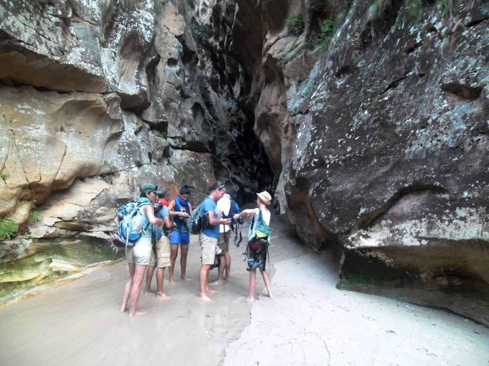 A group of barefoot hikers with rucksacks on damp sand at the entrance to a narrow passage between tall sandstone walls.