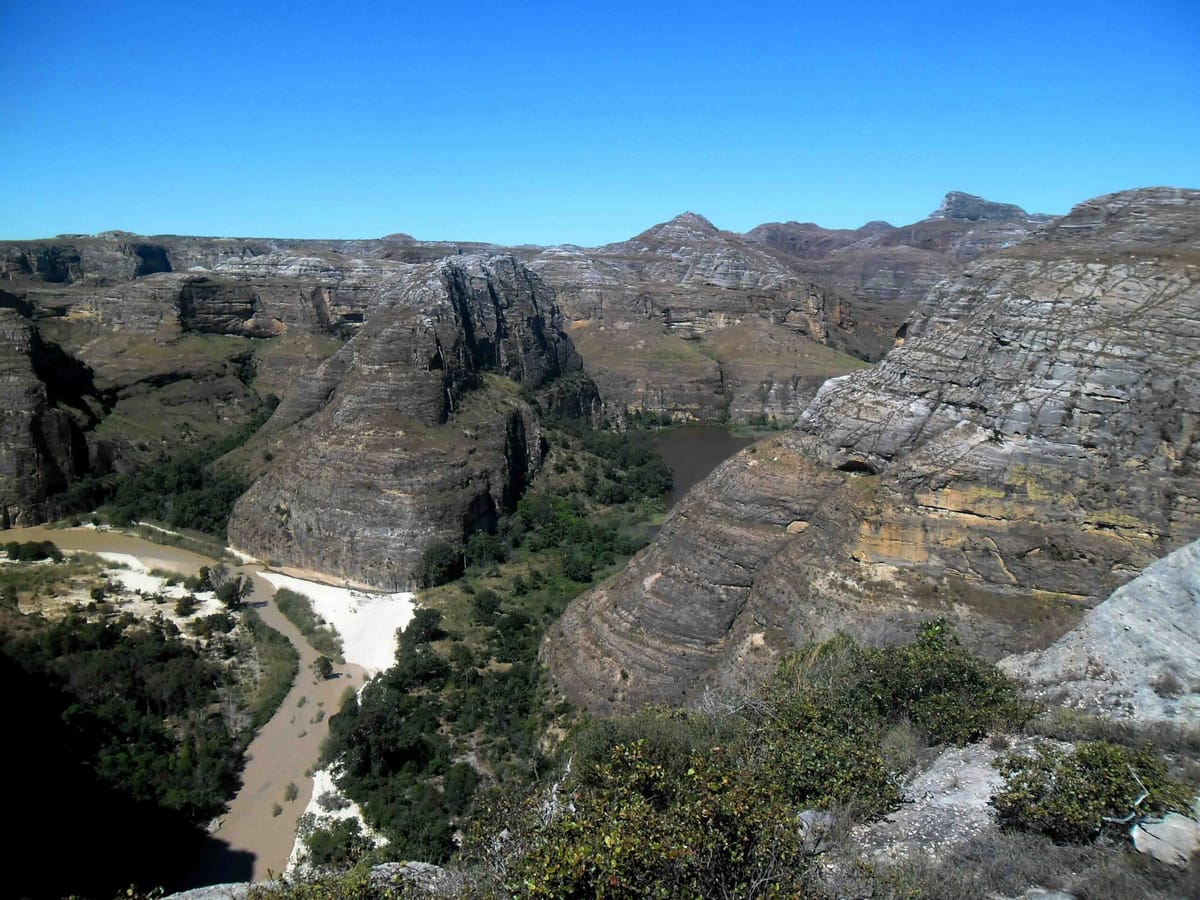 A sandstone canyon with streaked walls seen from above, with a river edged by white sandbanks winding between the cliffs and vegetation.