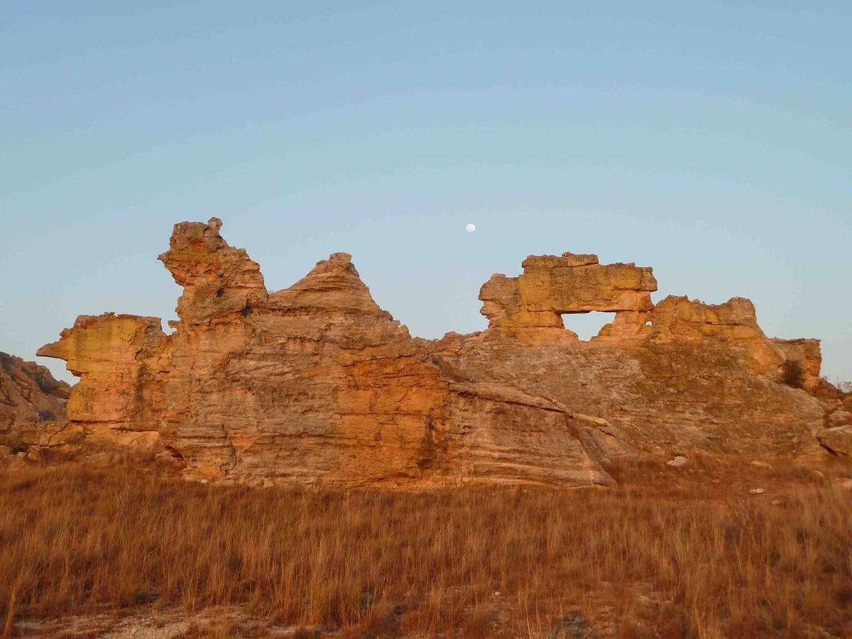 Weathered sandstone rocks pierced by a small arch, glowing gold in low light above a savannah of dry grass, with the moon already up.