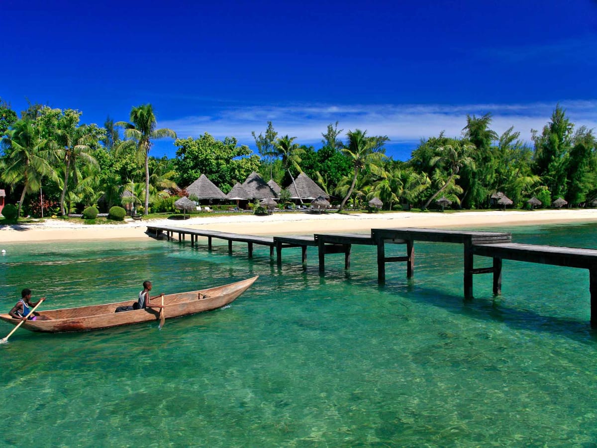 Two young men in a dugout canoe on the turquoise lagoon of Sainte-Marie, near a wooden jetty and bungalows.