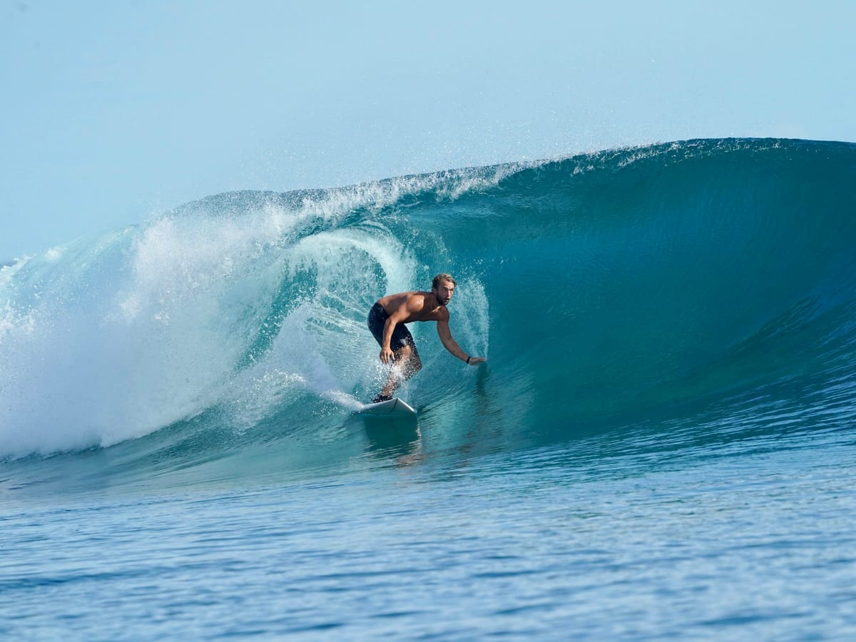 A bare-chested surfer gliding beneath the lip of a curling turquoise wave on a sunny day.