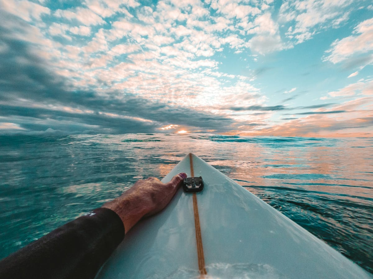 A wetsuit-clad surfer's hand resting on the nose of his board, facing out to sea under a sunset sky.