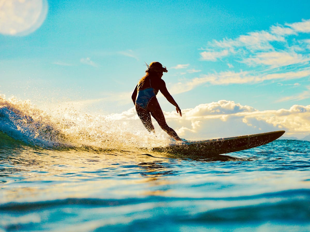 A surfer in a swimsuit standing on a longboard, gliding through a spray of golden foam beneath a blue sky with the moon just visible.