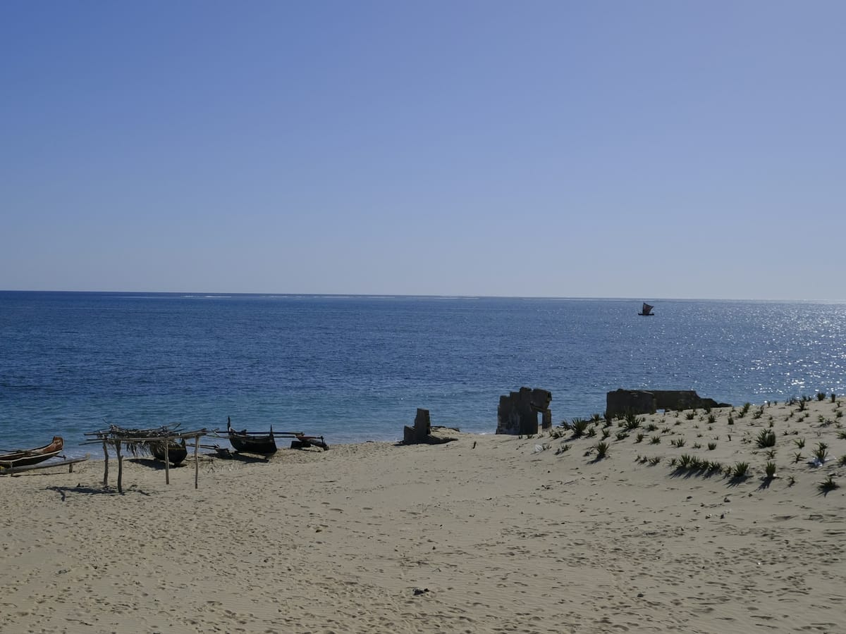 Outrigger canoes pulled up on a sandy beach at Sarodrano, near Toliara, facing a blue sea where a sailing pirogue passes by.