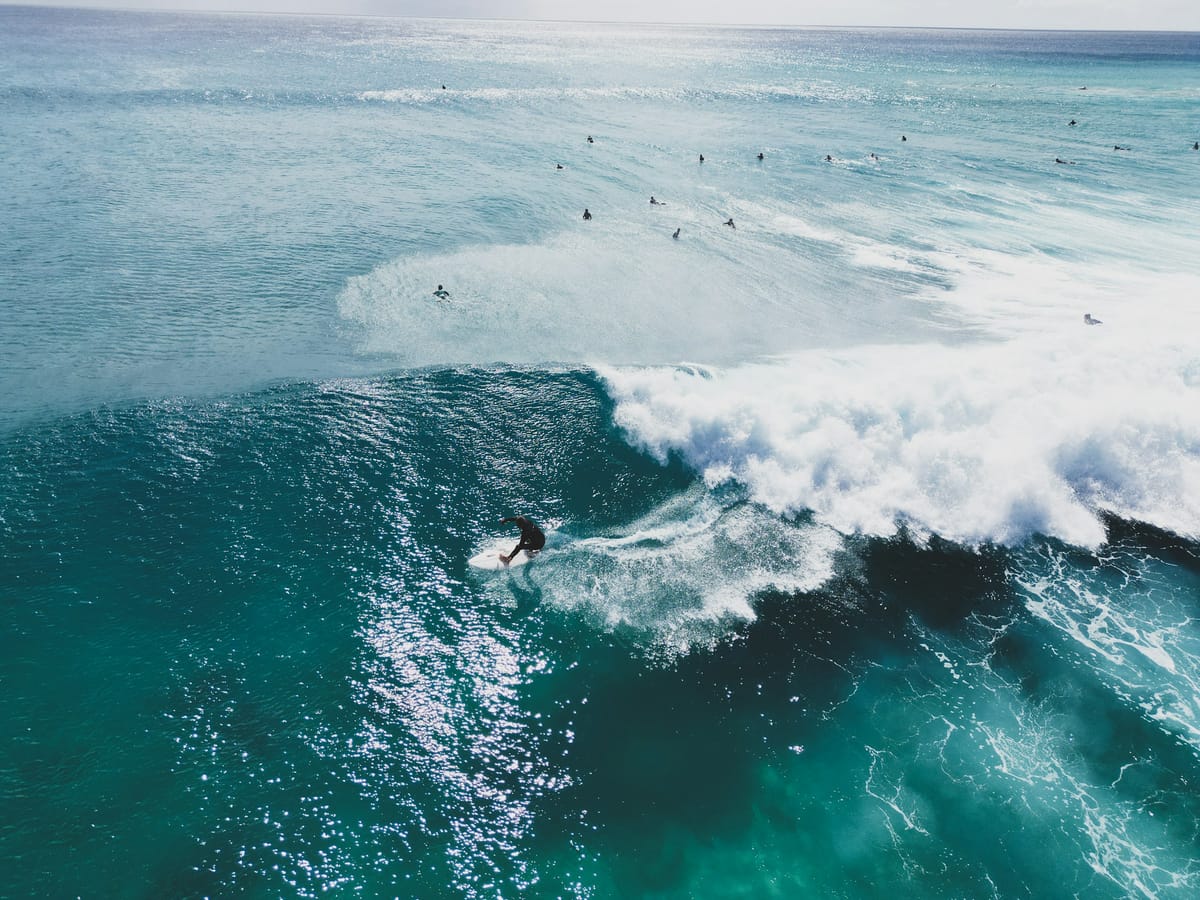 Aerial view of a surfer in a wetsuit turning at the foot of a wave breaking into white foam, while other surfers wait further out.