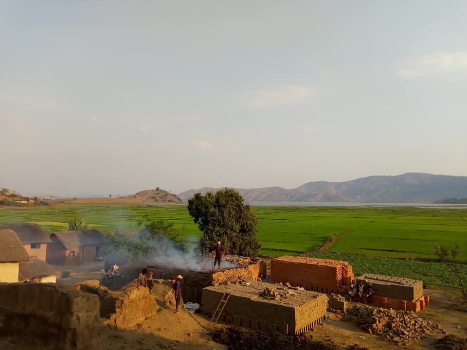 Smoking brick kiln, stacks of red bricks and earthen houses in front of the green rice paddies of Talata Volonondry.